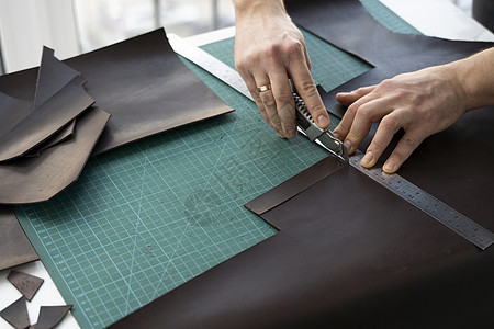 男人的手拿着文具刀和金属尺子 在他的车间里切割皮革钱包的碎片 与棕色天然皮革的工作过程 拿着制作工具的工匠制造业皮匠腰带皮艺机器图片