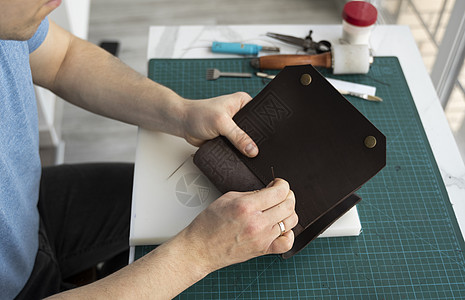 男士手在他的工作室里制作皮革钱包 与棕色天然皮革的工作过程 拿着制作工具的工匠拳头皮匠缝纫锥子皮肤维修剪刀材料鞋类产品图片