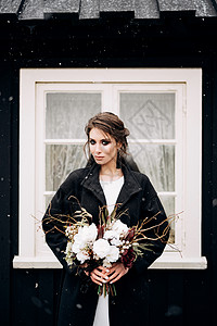 新娘身穿白色丝绸婚纱和黑色外套的肖像 手里拿着新娘的花束 有一个白色窗口的黑木房子 下雪 目的地冰岛婚礼牡丹头发薄纱化妆品裙子丝图片