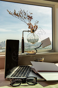 带笔记本电脑的工作区笔记本铅笔太阳镜咖啡杯窗边的干花花瓶可欣赏城市景观技术互联网窗户玻璃桌子键盘软垫反射文档办公室图片