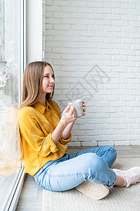 穿着黄色衬衫 坐在地上喝茶的有魅力的少女女子女性杯子饮料成人快乐窗户微笑闲暇咖啡幸福图片