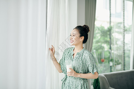 年轻微笑的女人从玻璃杯里喝牛奶的早晨画像 女人站在窗边的家里 吃着健康的食物图片