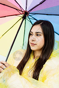 美丽的黑发美女 在雨中拿着多彩的雨伞公园乐趣天气女士微笑季节雨衣女性快乐彩虹图片