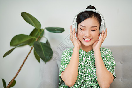 亚洲妇女在家里用耳机监听音乐 带复制空间的无线科技情人技术女士音乐播放器金发歌曲乐趣窗户沙发背景图片