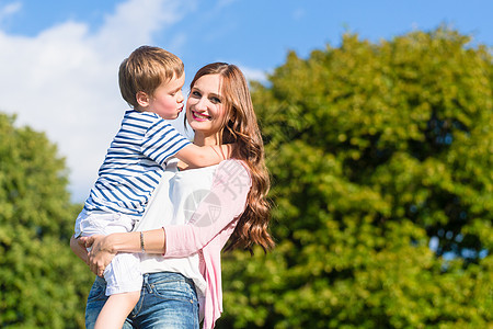 母亲抱着儿子的双臂亲吻他草地父母蓝色太阳幸福女士快乐公园妈妈乐趣图片