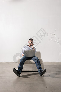 年轻人坐在空房间里 使用手机和笔记本电脑机构自由职业者产业男人沙发创始人企业家职场电话创意图片