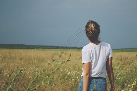 一个身穿白色 T 恤和牛仔裤的金发女孩站在田野里衣服场地蓝色天空女士金发女郎女性衬衫图片