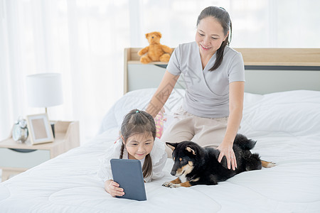 亚洲小女孩展示平板电脑 让她的黑什叶派狗感兴趣并和母亲一起睡在白床上 幸福家庭的概念与自己的宠物呆在家里会感到轻松图片