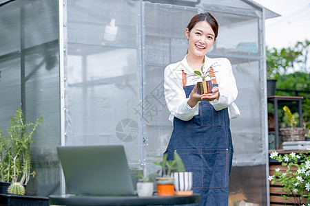 美丽的亚洲女孩还站在温室工作场所的前面 持有植物产品并展示给摄像头看图片