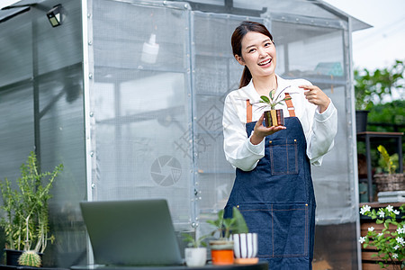 美丽的亚洲女孩拿着一盆植物 指着一盆植物 还看着站在她家前面绿色花园里的相机 小型企业的可持续发展与种植植物的概念有关图片