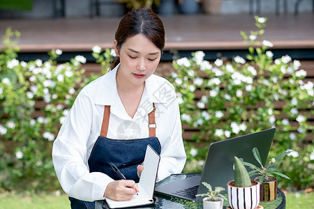 美丽的亚洲女人将数据记录到笔记本电脑前的笔记本中 并留在家里的花园里 小型企业的可持续发展与种植植物的概念有关图片