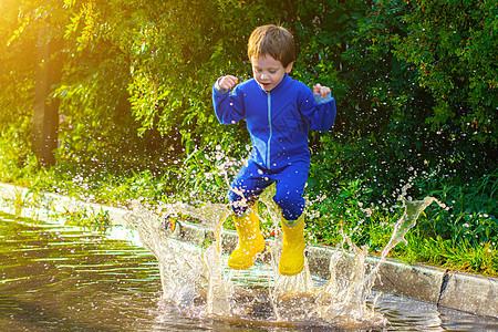 一个穿着橡胶靴的快乐男孩在水坑里跳跃 男孩跳进水坑里 坏天气 雨后的水坑 一个穿着橡胶靴的孩子 夏日散步 快乐的孩子雨靴彩虹家庭图片