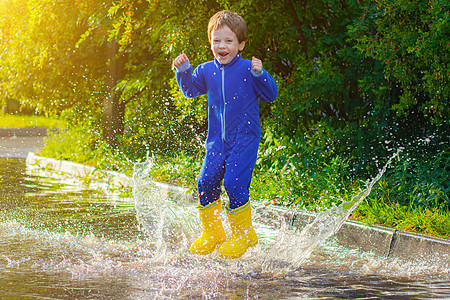 一个穿着橡胶靴的快乐男孩在水坑里跳跃 男孩跳进水坑里 坏天气 雨后的水坑 一个穿着橡胶靴的孩子 夏日散步 快乐的孩子乐趣下雨公园图片