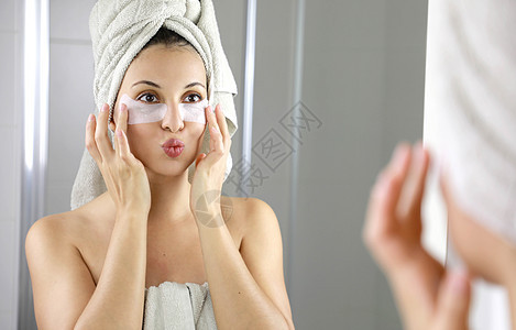 美丽的女人在浴室的镜子里亲吻自己 皮肤照护的女孩触摸眼睛下的布面罩以减少眼袋;她用脸巾遮住她的头部图片