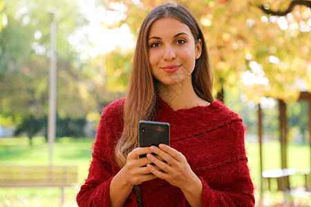 时装女郎拿着手机看秋天市公园的摄影机 穿着红色红袍图片