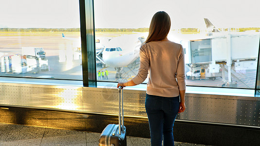 机场的年轻女子透过窗户看飞机航空女士建筑天空通勤者时间假期航班行李飞机场图片