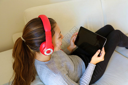 快乐的年轻女子选择在平板电脑上的音轨 同时躺在沙发上 在阁楼公寓里用耳机戴耳机耳塞房间药片窗户音乐玩家闲暇阁楼微笑长椅图片