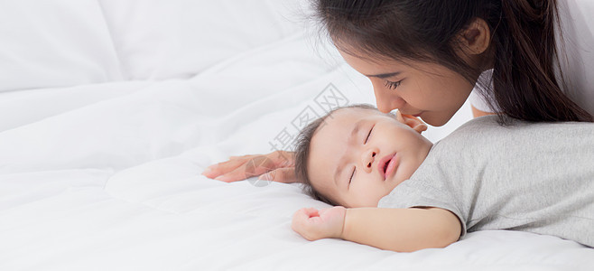 年轻的亚洲妈妈在卧室的床上亲吻小女孩的脸颊 妈妈爱新生儿和照顾 妈妈和孩子一起表达 父母和女儿 家庭观念童年女性婴儿投标成人母性图片