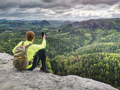 跑轨者休息 用他的智能手机拍照黄绿色游客背包男人悬崖享受运动鞋照片齿轮探索图片