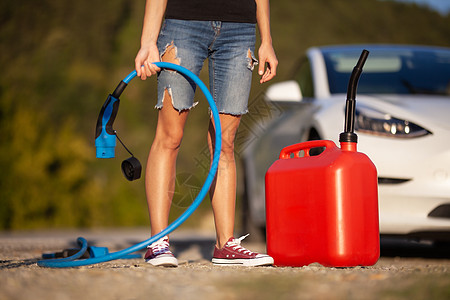 女孩站在电动汽车旁边 持有充电电缆和煤气罐头图片