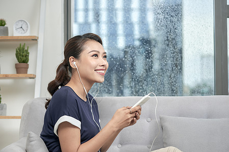 快乐的年轻亚洲女人戴着耳机在家里用智能手机发消息 听音乐 舒适地坐在灰色沙发上女士女性白色沙发成人闲暇长椅电话图片