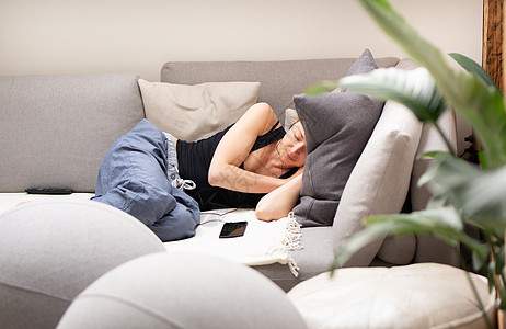 坐在舒适的沙发上睡午觉的疲劳年轻女子 在客厅里坐着大垫子 她旁边有一部手机图片