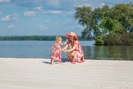 一个穿着浅色夏日太阳裙的迷人女孩和她的小女儿在沙滩上散步 享受温暖阳光明媚的夏日乐趣海滩微笑婴儿女士天空家庭幸福童年孩子图片