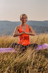 美丽的女人坐在莲花的姿势 用耳机和冥想在自然中成人训练天空草地生活方式瑜伽灵活性沉思教练力量图片