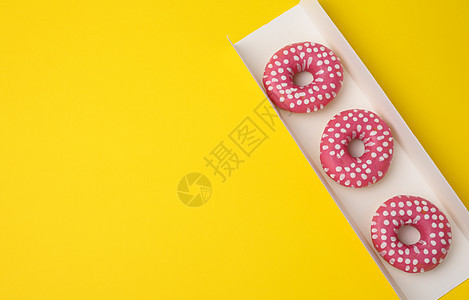 三个烤圆甜甜圈 粉红色釉料 放在黄色背景的白色纸箱里 顶视图图片