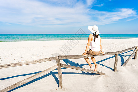 独自度假的年轻女子在蓝天下的白色沙滩上坐在木栅栏上欣赏清澈透明的热带海洋 戴着大白帽享受夏季旅行的沉思女孩图片