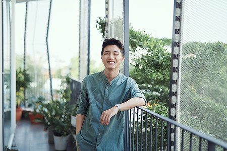 年轻的亚洲人等待和靠在手扶栏杆上男性环境阳台植物微笑男人享受爱好背景