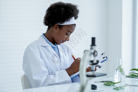年轻的非裔美国科学家记录分析数据 并与实验室的其他工具一起坐在房间里 并用植物组织科学概念进行实验图片