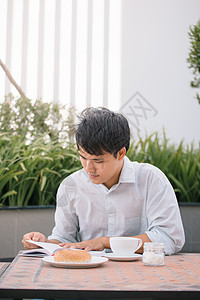 一个英俊男人的肖像 阅读一本书 享受他的咖啡白色桌子早餐房间杯子成人食物商业面包图片