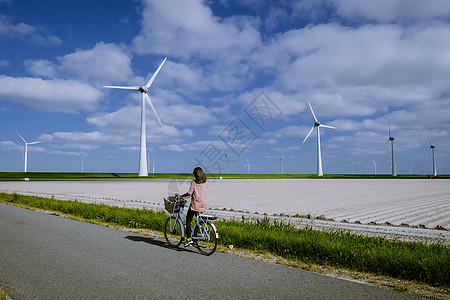 风车农场旁的年轻女性电动绿色自行车 风车在美丽明亮的日子里与世隔绝 荷兰植物生态公园能源全球技术发电机场地活力海洋图片