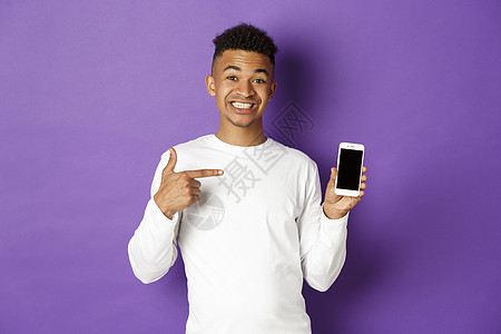 英俊的非洲美洲男子肖像 对智能手机屏幕微笑并指手指脚 展示应用程序 站在紫底背景上学生多样性工作紫色促销成人广告手势爆炸发型图片