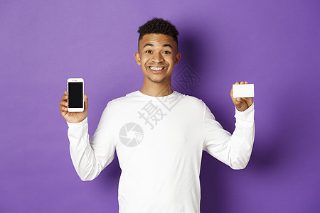 英俊的非洲美洲男子的肖像 微笑着喜悦 展示智能手机屏幕和信用卡 站在紫底背景之上快乐促销情感银行男性成人爆炸金融员工学生图片