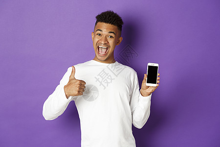 照片显示非洲男子穿着白色运动衫 微笑并举起拇指支持批准 展示手机屏幕 站在紫色背景上站立男性健康工作室促销发型购物网络技术学生情图片