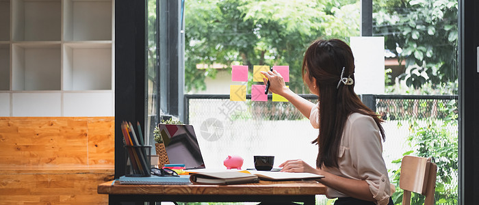 有商业魅力的年轻亚洲女性在便条纸上写笔记 安排与坐在后台的同事会面 透过玻璃拍照 创业和财务概念图片