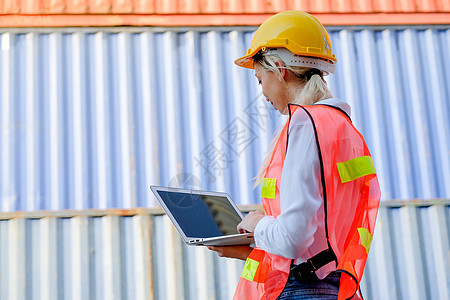 技术员的背部有妇女用笔记本电脑工作 站在日光灯照亮的货柜前商业拖拉机运输女士服务互联网仓库经理工程师技术图片