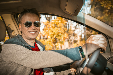 司机在秋林拦下汽车 太阳镜上的帅男对坐在车内 窗外树和黄色叶子背景的照相机微笑 脸朝着镜头笑图片