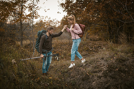 在大自然中远足 年轻男女背包客手拉着手从斜坡上下来 手里拿着登山杖 背着背包的游客穿过秋天的森林 支持理念图片
