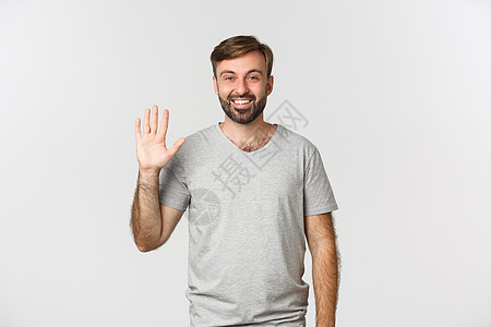 穿着灰色T恤的英俊caucasian男子肖像 向她问好 友好挥手 站在白背景上情绪微笑男性学生快乐成功工作室胡须广告胡子图片
