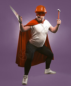 拿着建筑工具的超级英雄人 红色头盔的工程师或修理工和超级英雄制服 锯子和锤子站在葡萄紫色背景中图片