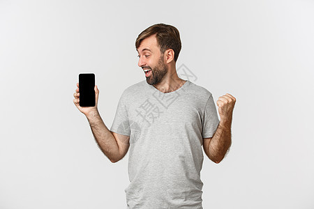 兴奋的留胡子男子肖像 在应用程序中达到目标 展示移动电话屏幕和欢乐 赢得东西 站在白背景上站立商业衬衫互联网情绪化网络快乐消息情图片