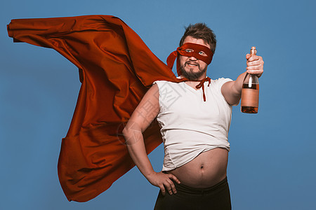 喝醉的超级英雄或反英雄拿着一瓶酒 戴着超级英雄红色面具 戴着飞行斗篷的男人拿着一瓶酒 同时露出大肚子 在褪色的牛仔蓝色背景中被隔图片