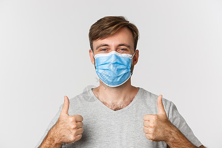 流行病 covid19 和社会疏远的概念 戴着医用面具的快乐白人男子的特写 竖起大拇指 站在白色背景上疾病促销标识口罩流感男性衬图片