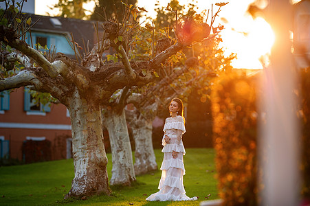 穿着白婚礼服的新娘 位于奥地利城镇的一个公园中 日落时有大树长发婚礼女士裙子压痛形象建筑学村庄青年魅力图片