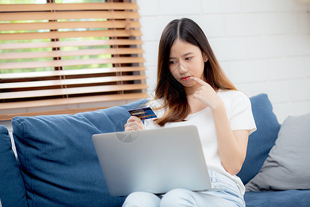 年轻的亚洲女性坐着思考想法 使用信用卡和沙发上的笔记本电脑 女孩在线购物购买和支付 沙发上的笔记本 金融和借记卡 生活方式概念商图片