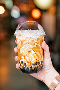 年轻女孩在台湾背景的夜市里 拿着并展示一杯咖啡棕色糖味木薯珍珠泡泡奶茶 闭紧了 bokeh牛奶珍珠波霸杯子塑料老虎市场旅行斑纹食背景图片