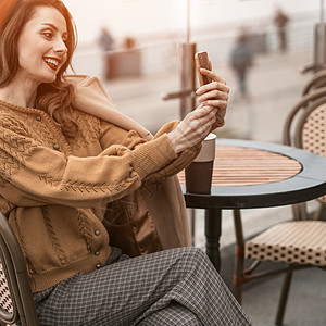美丽的年轻女子坐在春天的露天咖啡馆里一边喝着咖啡一边用她的智能手机进行视频通话 红色贝雷帽的法国妇女有都市背景的 正方形裁剪时尚背景图片